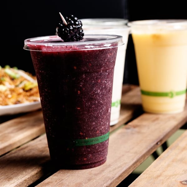 A dark berry smoothie with a blackberry garnish, alongside other drinks and food on a wooden table.