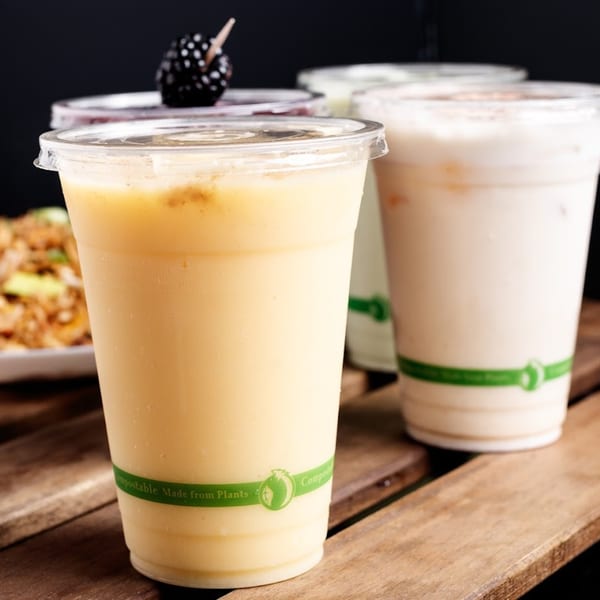 Four healthy Nokturne World Kitchen & Juice smoothies in compostable cups on a wooden table.