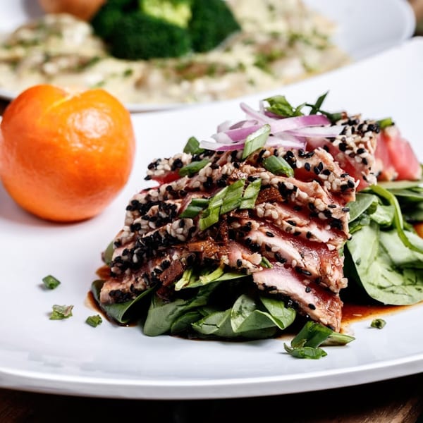 Seared tuna with sesame seeds, red onion, and spinach, next to an orange on a white plate.