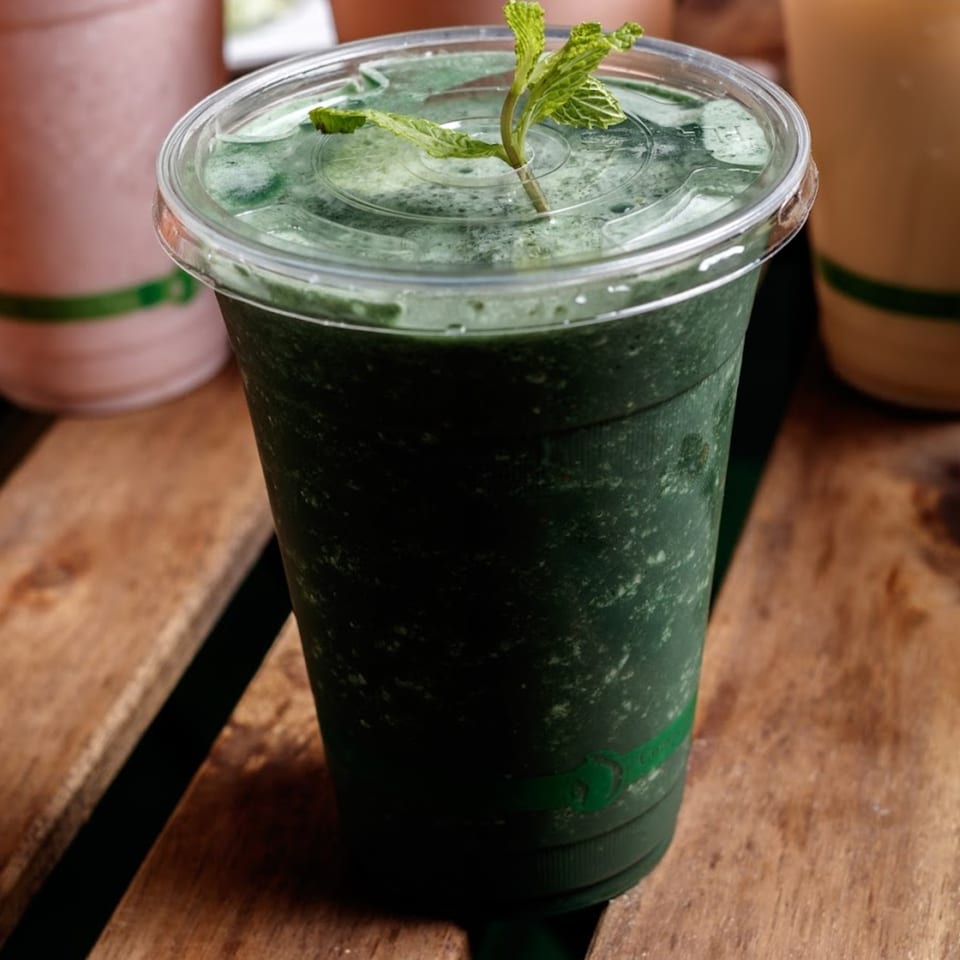 A vibrant green smoothie with a mint garnish in a clear cup on a rustic wooden surface.