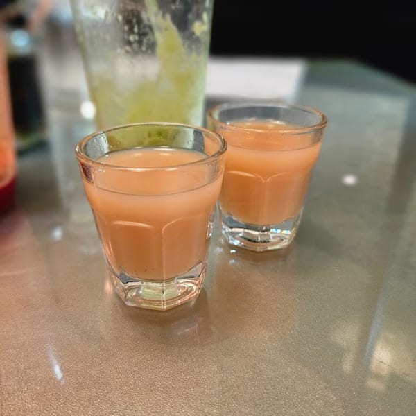 Two small glasses of orange juice shots on a table, with a blurred green drink in the background.