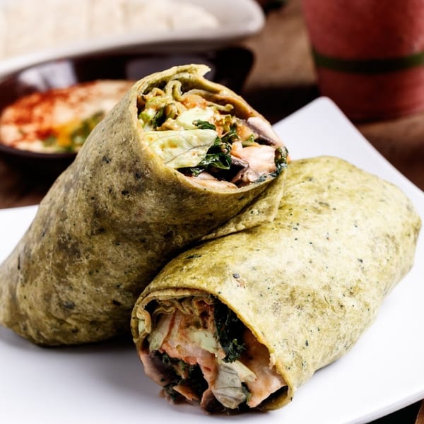 Two healthy wraps with salmon, kale, and mushrooms on a white plate, with hummus in the background.
